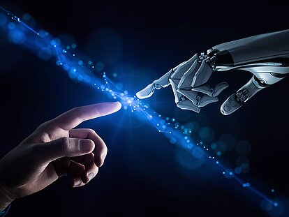 Kuenstliche Intelligenz, nach Da Vinci KI-Darstellung, menschlicher und künstlicher Finger treffen sichvor einem dunklen Hintergrund und erzeugen einen schräg von links oben nach rechts unten verlaufenden blau leuchtenden Lichtstrahl.